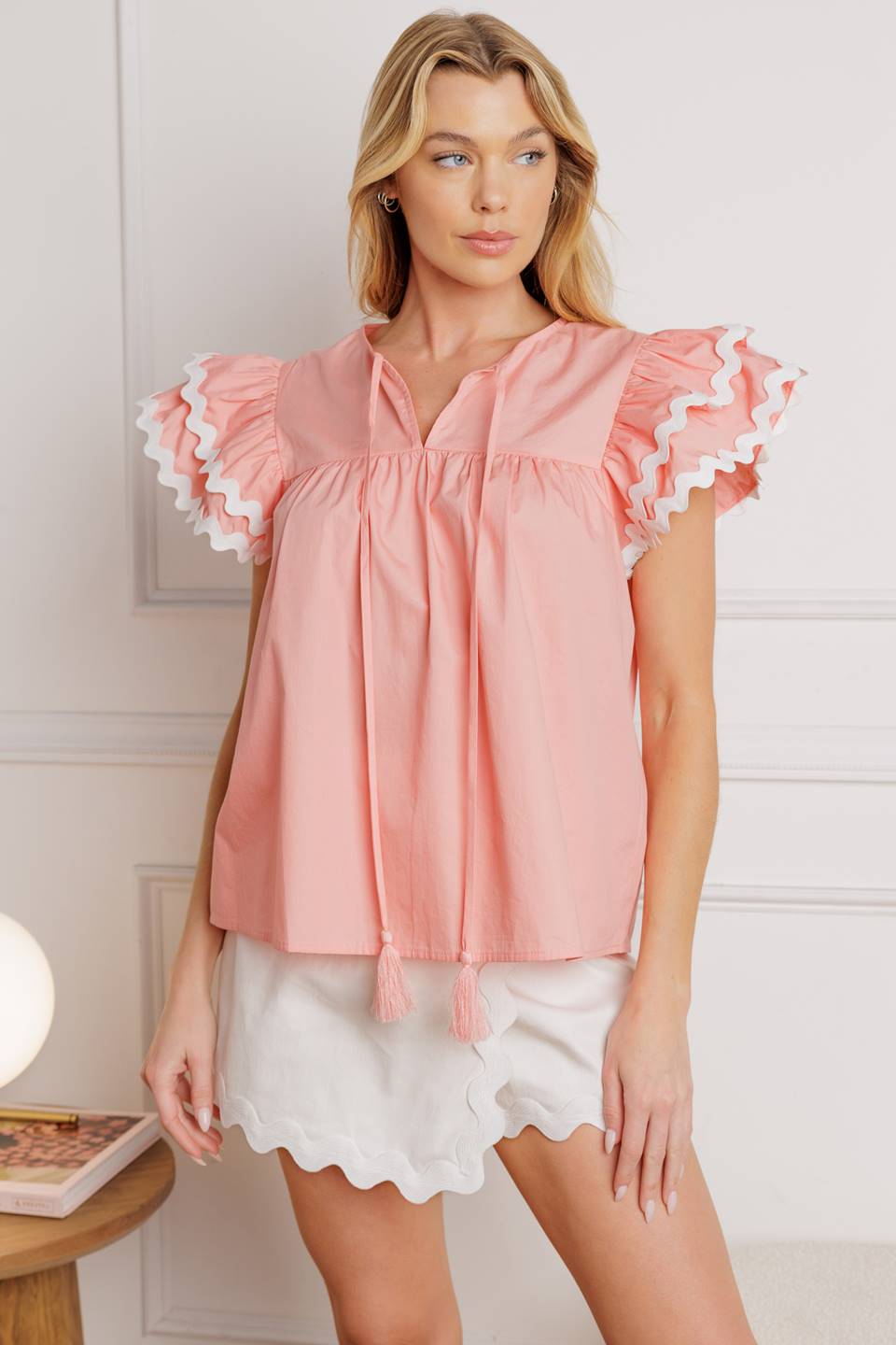 A woven top with a round neckline, tie and tassel, and double ruffled sleeves with ric rac trimming.